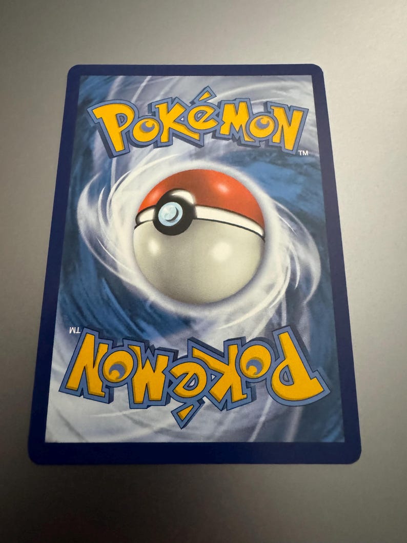 Puede incluir: El reverso de una carta coleccionable de Pok&eacute;mon. Muestra el logotipo de Pok&eacute;mon en amarillo y azul, y un dise&ntilde;o de Pok&eacute; Ball en el centro. La tarjeta tiene un borde azul y un fondo arremolinado.