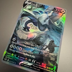 May include: A holographic Lugia V Pokémon trading card. The card features a detailed illustration of Lugia, a large, white and blue Pokémon, with the text "Lugia V" at the top. The card has various game instructions and a HP of 220.
