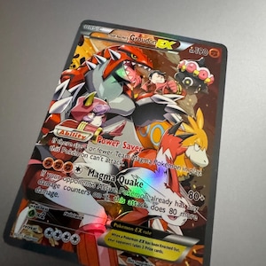 May include: A holographic Pokémon trading card featuring Groudon EX. The card has a red, orange, and white color scheme with various characters and text. The card's text includes "Magma Quake" and "Power Saver".