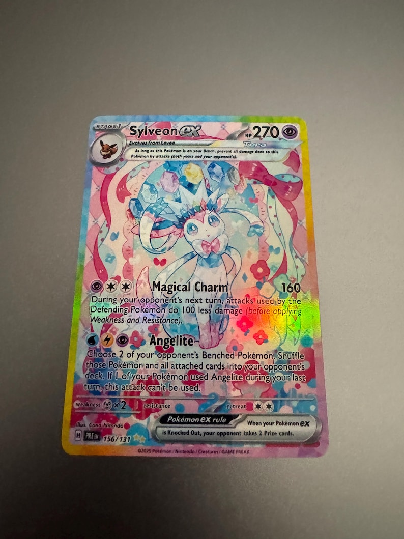 May include: A holographic Pok&eacute;mon trading card featuring Sylveon EX. The card has a colorful design with pink, blue, and yellow accents. Text includes "Magical Charm" and "Angelite" abilities. The card is part of the 156/131 set.
