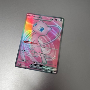 May include: A holographic Pokémon trading card featuring Mew ex. The card has a pink and iridescent design with the text "Mew ex" at the top and "Genome Hacking" below the image of Mew. The card is a collectible item.