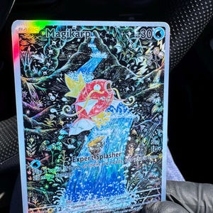 May include: A holographic Magikarp Pokémon trading card. The card features a red and yellow Magikarp leaping from a waterfall. The card has the text "Magikarp" and "Expert Splasher". The card is from the Paldea Evolved set.
