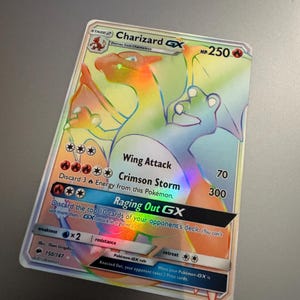 May include: A holographic Charizard GX Pokémon card. The card features a rainbow-colored illustration of Charizard with the text "Wing Attack", "Crimson Storm", and "Raging Out GX". The card also has the HP of 250.