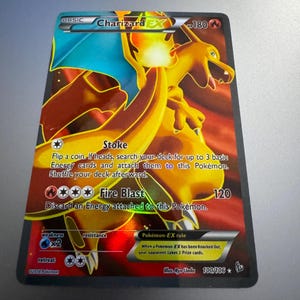 May include: A Charizard EX Pokémon trading card. The card features a vibrant illustration of Charizard in shades of orange, yellow, and blue. The card has text that includes "Stoke" and "Fire Blast".