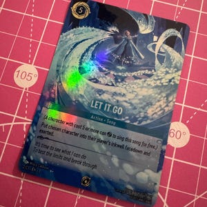May include: A collectible card with a blue and white design featuring a character in a flowing gown, with the text "LET IT GO". The card has a holographic effect and includes game instructions and a card number.