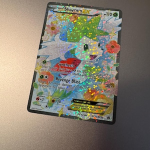 May include: A holographic Shaymin EX Pokémon card. The card features a Shaymin character with green and white coloring, surrounded by flowers. The card text includes "Synthesis" and "Revenge Blast" abilities, with a 110 HP rating.