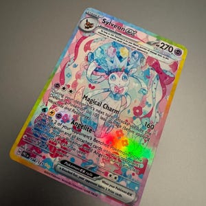 May include: A holographic Sylveon ex Pok&eacute;mon trading card. The card features a colorful illustration of Sylveon with pink, blue, and white accents. The card has text that includes the Pok&eacute;mon's name, HP, and attack details. The card is a collectible item.