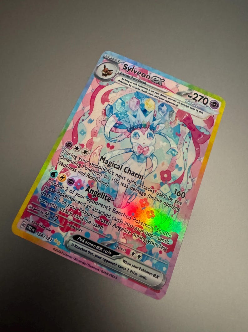 May include: A holographic Sylveon ex Pok&eacute;mon trading card. The card features a colorful illustration of Sylveon with pink, blue, and white accents. The card has text that includes the Pok&eacute;mon's name, HP, and attack details. The card is a collectible item.