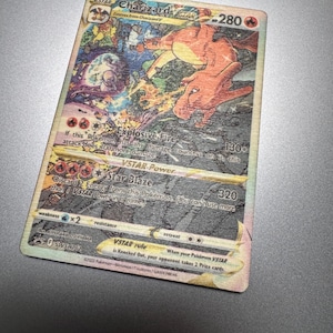 May include: A Charizard VSTAR Pokémon trading card. The card features a vibrant illustration of Charizard in action, with the text "Explosive Fire" and "Star Blaze" visible. The card has a holographic finish and is set against a gray background.