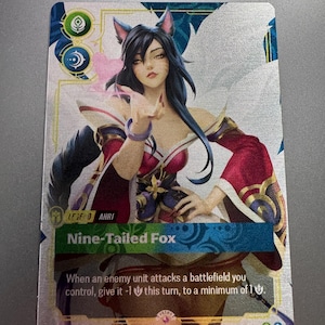 May include: A collectible trading card featuring an illustration of a dark-haired character with fox ears and tails. The card displays the text Nine-Tailed Fox, Ahri, and game mechanics text. The design includes red, blue, and gold accents on a metallic finish.