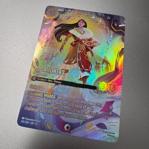 May include: A holographic trading card featuring Pocahontas from Disney Lorcana. The card showcases Pocahontas in a flowing outfit with text that reads "CALMING WORDS". The card has a shimmering, iridescent finish with a colorful background.