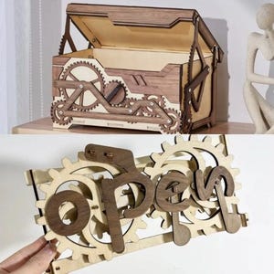 Può includere: Scatola di legno con coperchio incernierato e dettagli a forma di ingranaggio. La scatola è realizzata in legno marrone chiaro e scuro, con intricati disegni di ingranaggi sui lati e sulla parte anteriore. La parola "open" è visualizzata su un cartello di legno separato con dettagli di ingranaggi.