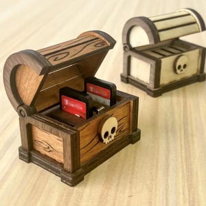 以下が含まれることがあります： ヒンジ付きの蓋が付いた、宝箱型の木製ボックスが2つ。 大きなチェストは開いており、赤と黒のゲームカートリッジが見えます。 両方のチェストはスカルデザインで、濃い茶色と明るい茶色の木材で作られています。