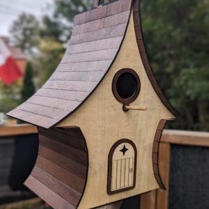 May include: Wooden birdhouse with a whimsical design. The birdhouse features a light brown body, a dark brown roof, and a small round entrance. A small wooden perch extends from the entrance. The birdhouse is mounted on a wooden fence.