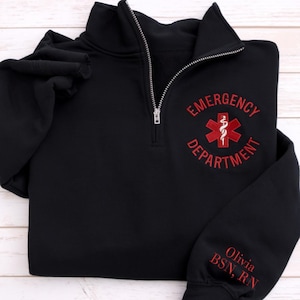 May include: Black quarter-zip sweatshirt with a silver zipper. The front features the words "EMERGENCY DEPARTMENT" embroidered in red, along with a red medical symbol. The sleeve has the name "Olivia BSN, RN" embroidered in red.