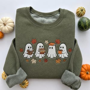 May include: An olive green crewneck sweatshirt featuring an embroidered design of four white ghosts. One ghost reads a book, one holds a stack of books, one has a book on its head, and one holds a mug, all surrounded by scattered orange and brown autumn leaves.