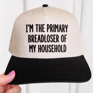 May include: A beige and black baseball cap with the text "I'M THE PRIMARY BREADLOSER OF MY HOUSEHOLD" in black. The cap has a flat brim and a button on top. The cap is being held by a person.