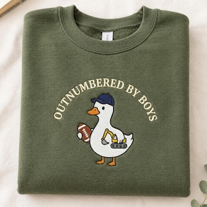 May include: Olive green sweatshirt with the text "OUTNUMBERED BY BOYS". The sweatshirt features a cartoon duck wearing a baseball cap and holding a football and a construction vehicle.