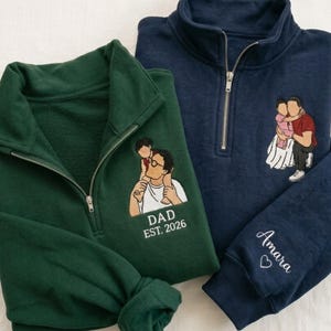 May include: Two embroidered quarter-zip pullovers. One is dark green with a graphic of a father and child, and the text "DAD EST. 2026". The other is navy blue with a family graphic and the name "Amara" on the sleeve.