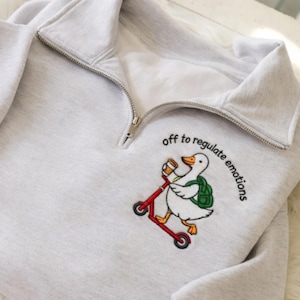 May include: A light gray quarter-zip sweatshirt featuring an embroidered graphic of a white duck riding a red scooter while wearing a green backpack and holding a coffee cup, with the text "off to regulate emotions" arched above the duck.
