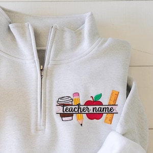 May include: A light gray quarter-zip sweatshirt featuring an embroidered design on the left chest. The graphic includes a coffee cup, a yellow pencil, a red apple, and an orange ruler, with the text teacher name printed in black script.
