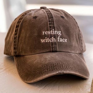 May include: A brown baseball cap with the phrase "resting witch face" embroidered in white. The cap has a curved brim and a distressed look, suggesting a vintage or worn style. The hat is sitting on a light-colored surface.