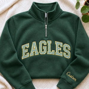 May include: A dark green quarter-zip sweatshirt featuring the word EAGLES in a white and green gingham pattern with a yellow outline across the chest. The word Custom is embroidered in yellow cursive script on the left sleeve cuff.