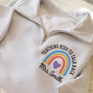 May include: Light gray sweatshirt with a quarter-zip collar. The sweatshirt features a rainbow design with the text "TEACHING KIDS TO TALK BACK" and "Mrs. Smith" in a circular arrangement.