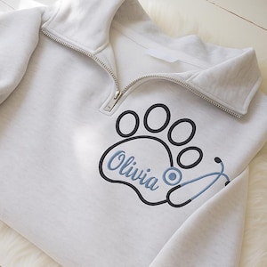 May include: A light gray sweatshirt with a quarter-zip collar. The design features a black paw print with the name "Olivia" in blue script, and a blue stethoscope. The sweatshirt is made of a soft material.
