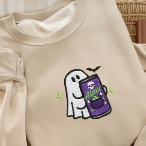 May include: Beige sweatshirt with a Halloween-themed embroidered design. The design features a white ghost holding a purple can with a skull and cauldron, the text "Alani", and a black bat. Green stars accent the design.