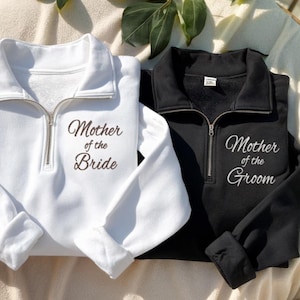 May include: A white quarter-zip sweatshirt with the text Mother of the Bride in brown and a black quarter-zip sweatshirt with the text Mother of the Groom in white. Both garments feature a fold-over collar and a silver-toned metal zipper.