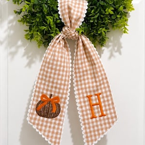 May include: A green boxwood wreath adorned with a hanging beige and white gingham ribbon. The ribbon features a scalloped white edge, an embroidered brown pumpkin with an orange bow on one tail, and an orange letter H on the other tail.