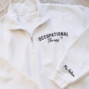 May include: White quarter-zip sweatshirt with the words "OCCUPATIONAL Therapy" and a heart embroidered in black. The sleeve has "Ms. Helen" embroidered in black. The sweatshirt has a silver zipper.