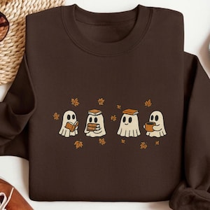 May include: A dark brown sweatshirt featuring a whimsical design of four cartoon ghosts. Each ghost is depicted with a different item: a book, multiple books, a book, and a mug. Small orange leaves are scattered around the ghosts.