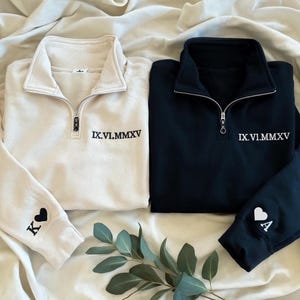 May include: Two personalized quarter-zip sweatshirts, one cream and one navy. Each features the date IX.VI.MMXV embroidered in white. The cream sweatshirt has a black heart and the letter "K" on the sleeve. The navy sweatshirt has a white heart and the letter "A".