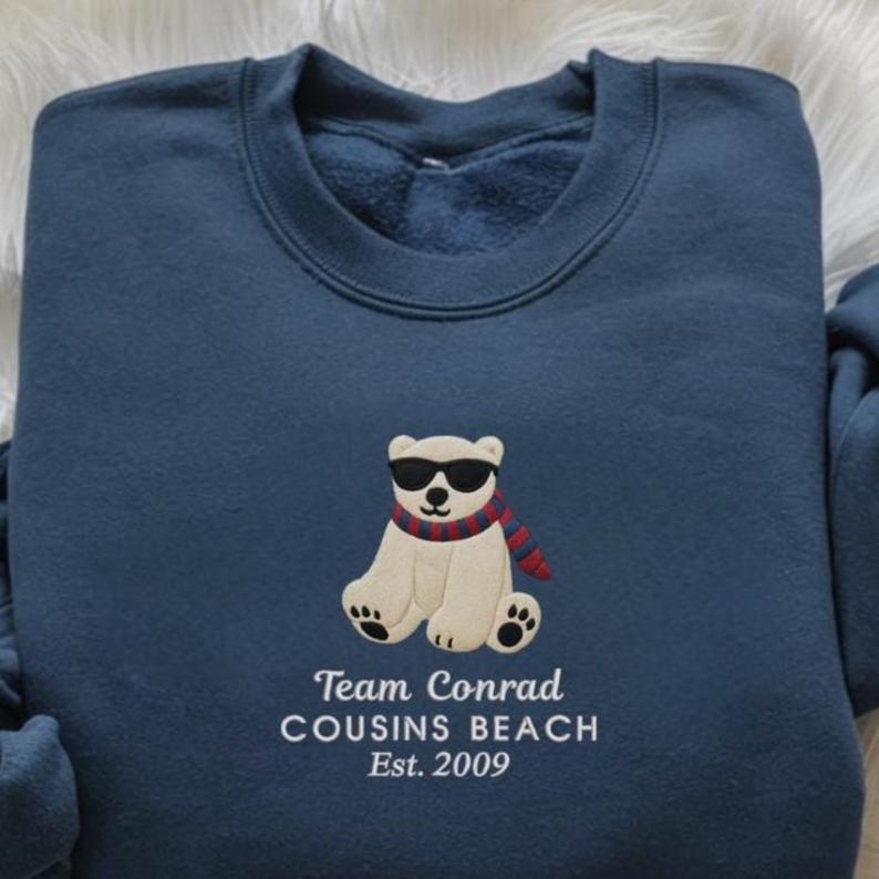 May include: Navy blue sweatshirt featuring an embroidered polar bear wearing sunglasses and a scarf. The text "Team Conrad Cousins Beach Est. 2009" is embroidered below the bear. The sweatshirt is made of a soft material.
