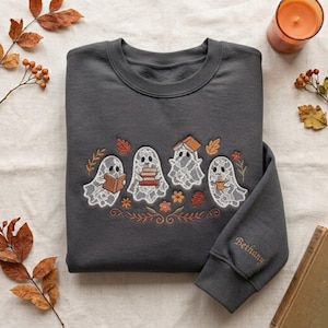 May include: A dark grey crewneck sweatshirt featuring an embroidered design of four white lace-patterned ghosts. One ghost holds a book, one balances a stack of books, one has a book on its head, and one holds a mug. The name Bethany is embroidered on the sleeve.