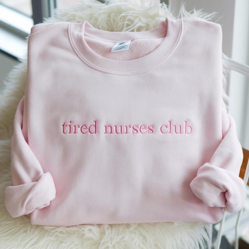 May include: A light pink sweatshirt with the embroidered text "tired nurses club" in a matching pink thread. The sweatshirt is folded, with the sleeves rolled up. The garment is displayed on a white, fluffy surface.