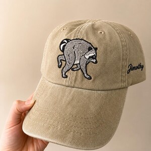 Peut inclure: Une casquette de baseball beige avec une broderie repr&eacute;sentant un raton laveur gris pench&eacute; sur le panneau avant. Le c&ocirc;t&eacute; de la casquette affiche le nom Jimothy brod&eacute; en &eacute;criture noire. La casquette poss&egrave;de une visi&egrave;re courb&eacute;e et des &oelig;illets.