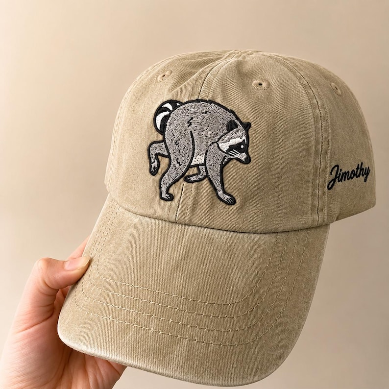 Peut inclure: Une casquette de baseball beige avec une broderie repr&eacute;sentant un raton laveur gris pench&eacute; sur le panneau avant. Le c&ocirc;t&eacute; de la casquette affiche le nom Jimothy brod&eacute; en &eacute;criture noire. La casquette poss&egrave;de une visi&egrave;re courb&eacute;e et des &oelig;illets.