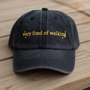 May include: A dark gray baseball cap with the embroidered text "very fond of walking" in gold. The cap has a curved brim and a button on top. The hat is made of a soft, worn material.