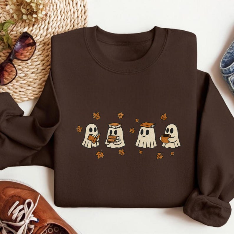 May include: A dark brown sweatshirt featuring a whimsical design of four cartoon ghosts. Each ghost is depicted with a different item: a book, multiple books, a book, and a mug. Small orange leaves are scattered around the ghosts.
