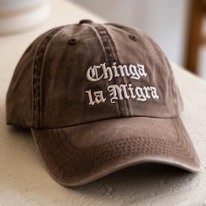 May include: Brown baseball cap with the embroidered text "Chinga la Migra" in white. The cap has a curved brim and a classic six-panel design. The hat is made of a soft, worn material.