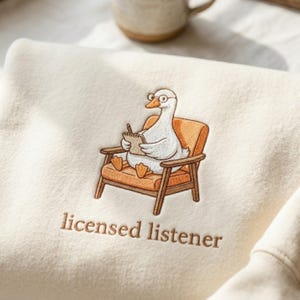 May include: Cream-colored sweatshirt featuring an embroidered cartoon goose wearing glasses and seated in an orange chair, holding a notepad. The text "licensed listener" is embroidered below the goose.