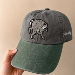 Peut inclure: Une casquette de baseball gris anthracite avec une visi&egrave;re verte, orn&eacute;e d'une broderie repr&eacute;sentant un raton laveur pench&eacute; sur le panneau avant. Le nom Jimothy est brod&eacute; en &eacute;criture cursive blanche sur le c&ocirc;t&eacute; de la casquette, dot&eacute;e d'&oelig;illets verts.