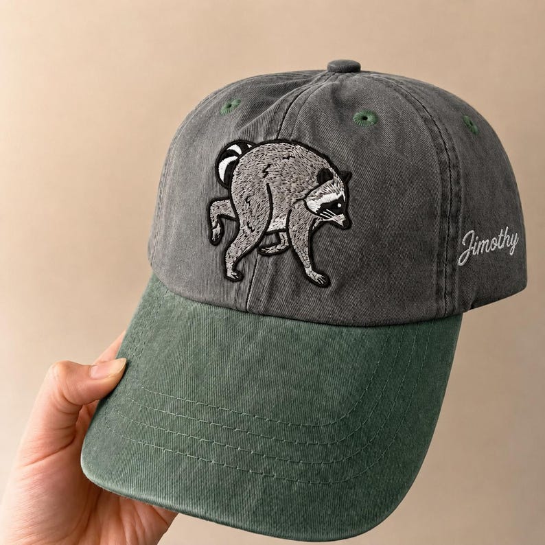 Peut inclure: Une casquette de baseball gris anthracite avec une visi&egrave;re verte, orn&eacute;e d'une broderie repr&eacute;sentant un raton laveur pench&eacute; sur le panneau avant. Le nom Jimothy est brod&eacute; en &eacute;criture cursive blanche sur le c&ocirc;t&eacute; de la casquette, dot&eacute;e d'&oelig;illets verts.