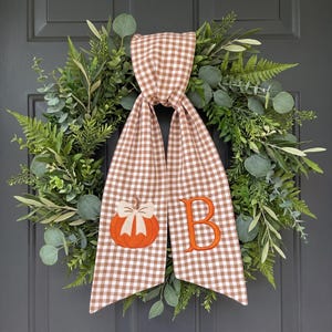 May include: A circular wreath composed of various green foliage including eucalyptus and ferns, featuring a long, brown and white gingham fabric ribbon draped over the center. The ribbon displays an embroidered orange pumpkin and the letter B.