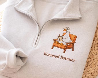 May include: A light gray quarter-zip sweatshirt featuring an embroidered graphic of a white goose wearing glasses, sitting in an orange chair while holding a notepad and pen, with the brown text "licensed listener" embroidered below the graphic.