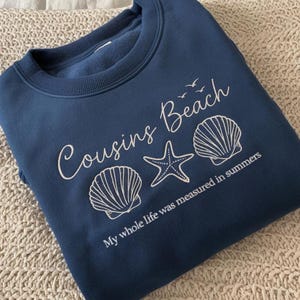 May include: A navy blue sweatshirt with the words "Cousins Beach" in white script, along with seashell and starfish graphics. The phrase "My whole life was measured in summers" is printed below the graphics.
