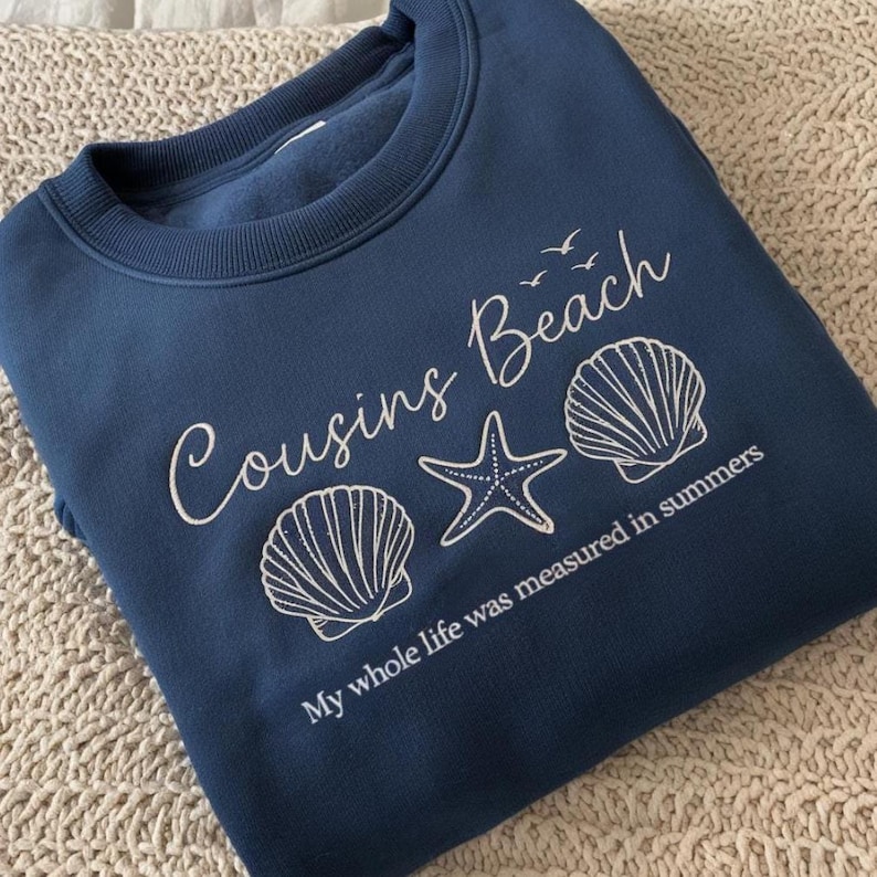 May include: A navy blue sweatshirt with the words "Cousins Beach" in white script, along with seashell and starfish graphics. The phrase "My whole life was measured in summers" is printed below the graphics.